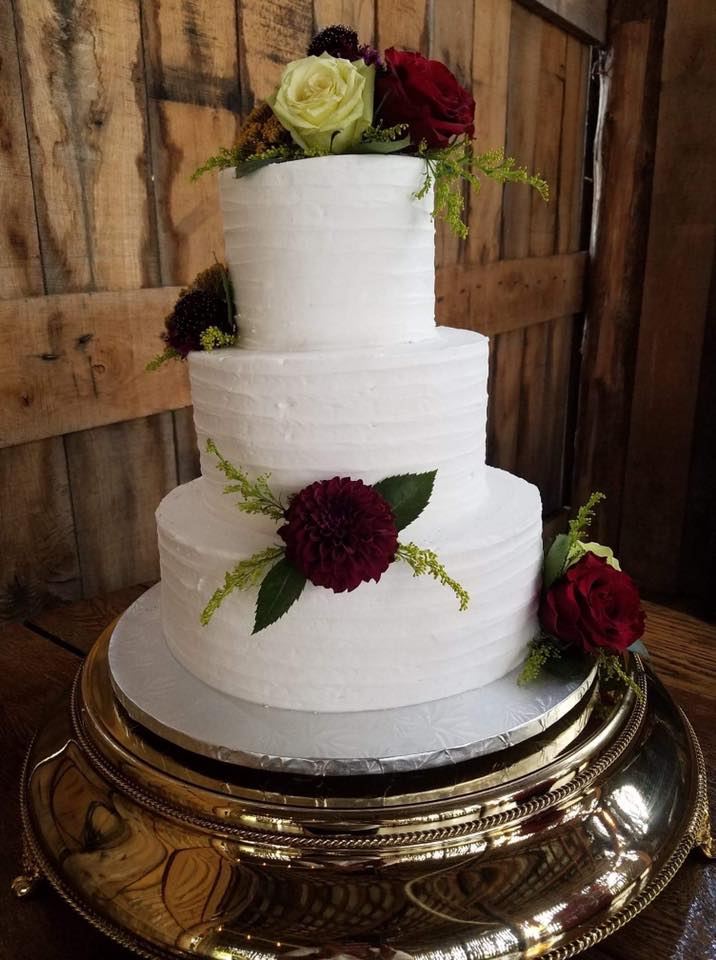A three-tier white wedding cake with burgundy roses on a gold stand